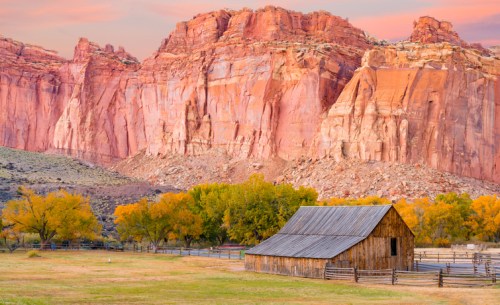 A rustic barn sits in a valley surrounded by vibrant autumn trees and towering red rock cliffs at sunset.