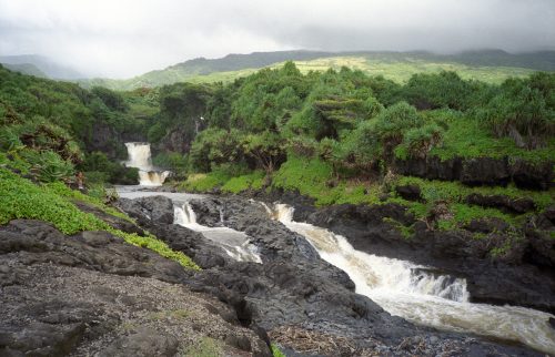 A lush landscape featuring cascading waterfalls and rocky terrain, surrounded by greenery under a cloudy sky.