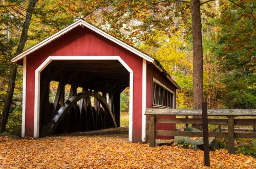A red covered bridge surrounded by autumn foliage and fallen leaves, with a wooden fence nearby.