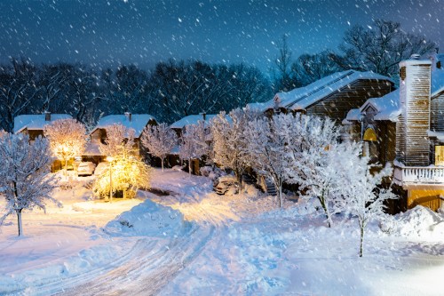 A snowy night scene with illuminated trees and houses, surrounded by a blanket of fresh snow and falling snowflakes.