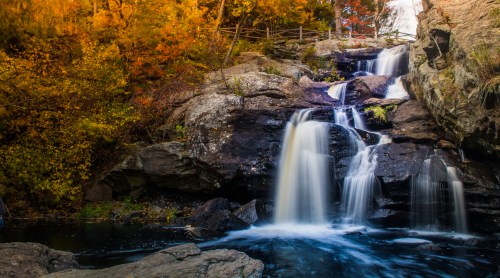 A serene waterfall cascades over rocks, surrounded by vibrant autumn foliage and a tranquil pool below.