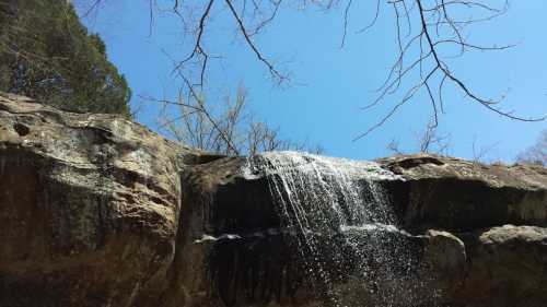 A waterfall cascades over a rocky ledge, surrounded by trees under a clear blue sky.