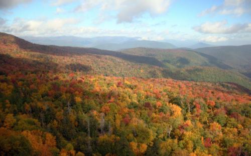 A panoramic view of vibrant autumn foliage covering rolling hills under a partly cloudy sky.