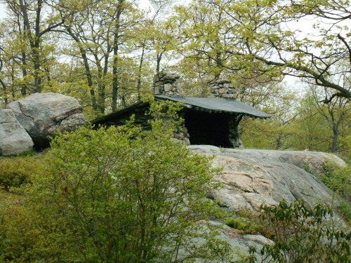 A small stone shelter with a sloped roof, nestled among large rocks and surrounded by greenery and trees.