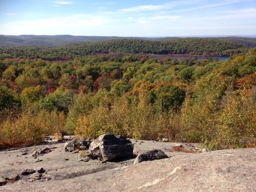 A scenic view from a rocky overlook, showcasing vibrant autumn foliage and a distant lake surrounded by hills.