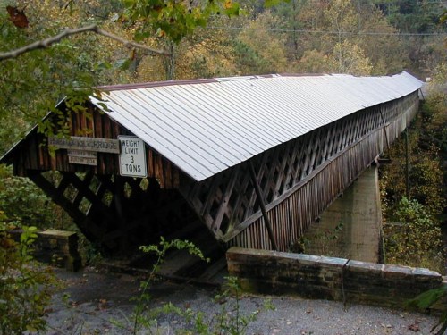 A wooden covered bridge with a sloped roof, surrounded by trees and autumn foliage, featuring a weight limit sign.