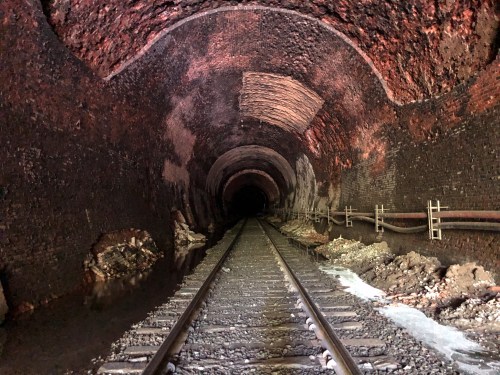 A dark, abandoned railway tunnel with brick walls and tracks leading into the distance, surrounded by debris and water.