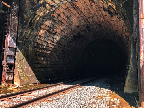 A dark tunnel entrance with stone walls and railway tracks leading into it, surrounded by graffiti and gravel.