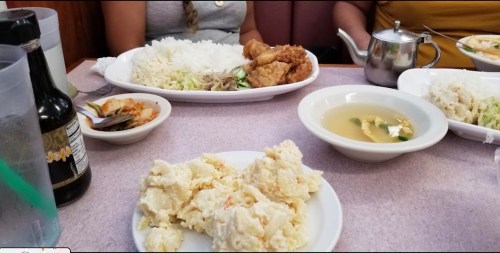 A table with plates of rice, fried chicken, salad, soup, and a side of potato salad, with drinks in the background.