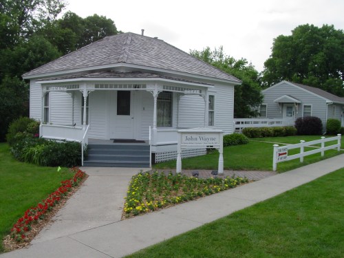 A white house with a porch, surrounded by a garden of flowers, featuring a sign that reads "John Wayne."