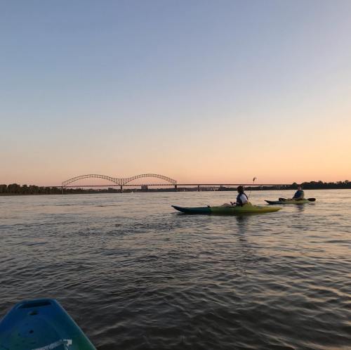 Two kayakers paddle on a calm river at sunset, with a bridge in the background against a colorful sky.