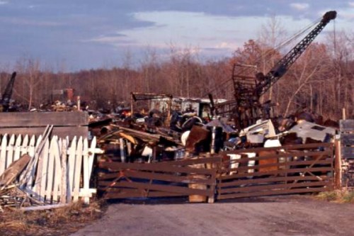 A cluttered junkyard with debris, old machinery, and a wooden fence under a cloudy sky.