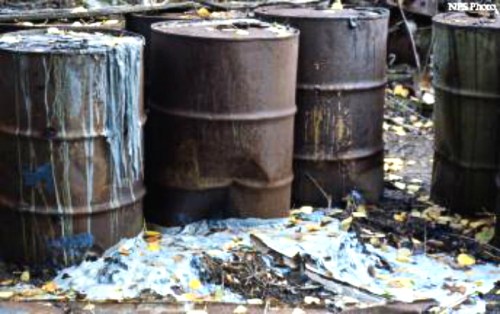 Rusty barrels with leaking liquid, surrounded by debris and fallen leaves in a neglected outdoor area.