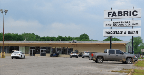 A fabric store with a large sign, featuring cars parked in front and a cloudy sky in the background.