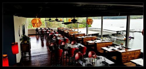 A spacious restaurant with wooden tables and red chairs, overlooking a lake with boats and large windows.