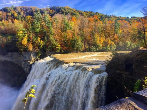 A waterfall cascades over a rocky ledge, surrounded by vibrant autumn foliage and a clear blue sky.