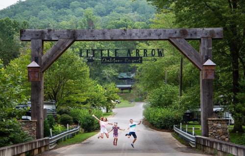 Three children jump joyfully under a wooden archway labeled "Little Arrow Outdoor Resort," surrounded by lush greenery.