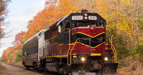 A vintage train with a red and black locomotive travels along a scenic route surrounded by colorful autumn foliage.