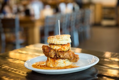 A plate with a stack of biscuits topped with fried chicken and drizzled with sauce, set in a casual dining environment.