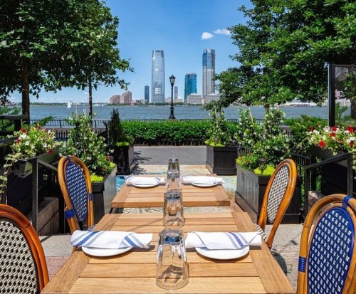 Outdoor dining setup with wooden tables and glasses, overlooking a waterfront and city skyline on a sunny day.