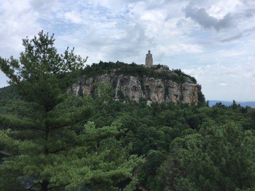 A rocky cliff topped with a tower, surrounded by lush green trees and a cloudy sky.