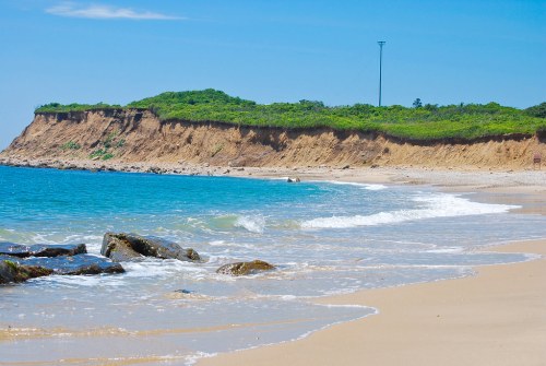 A serene beach scene with gentle waves, rocky shore, and a lush green cliff under a clear blue sky.