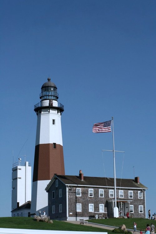 A tall lighthouse with a red and white design, next to a building, under a clear blue sky, with an American flag flying.