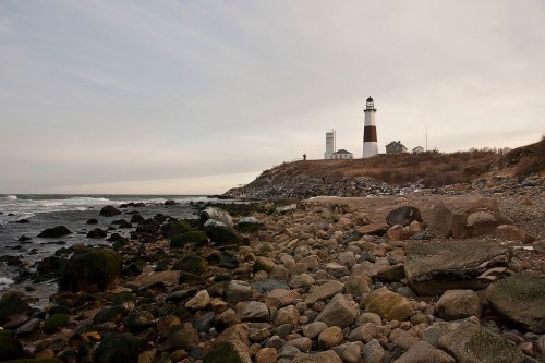 A rocky shoreline with a lighthouse on a hill, under a cloudy sky, near the ocean.
