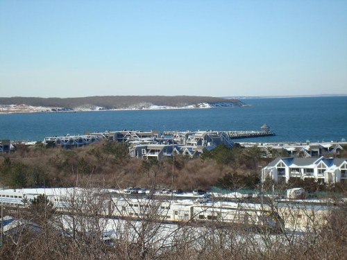 A coastal view featuring a snowy landscape, buildings near the shore, and a calm blue sea under a clear sky.