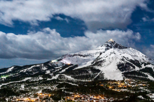 Snow-covered mountain under a night sky, with a small town illuminated at its base and clouds drifting above.