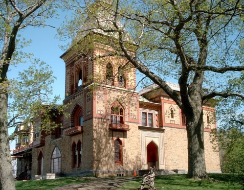 Historic building with intricate brickwork, tall tower, and large windows, surrounded by trees and green grass.