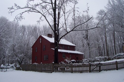A red house surrounded by snow-covered trees and a white yard, with a wooden fence in the foreground.