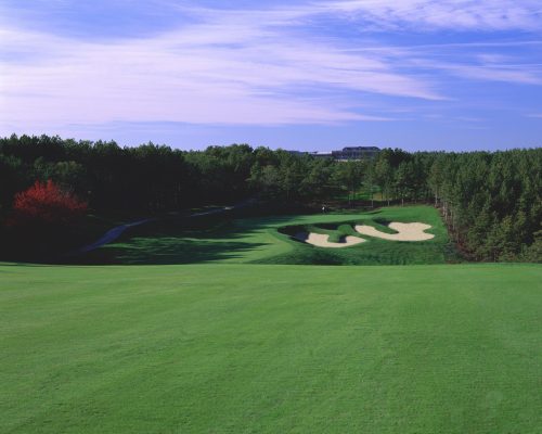 A scenic golf course with lush green grass, sand traps, and trees under a blue sky with wispy clouds.