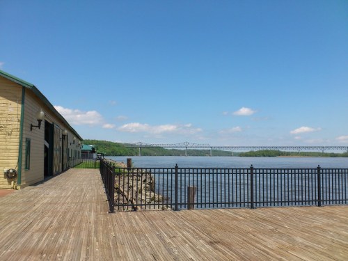 A wooden boardwalk along a river, with a bridge in the distance and clear blue skies above.