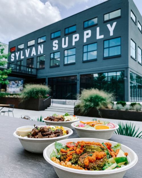 A variety of colorful bowls of food in front of a modern building with "SYLVAN SUPPLY" sign.