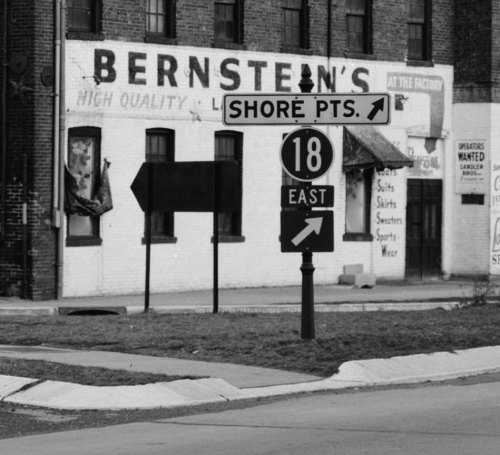 Black and white image of a street sign directing to Shore Points and indicating Route 18 East, with a building in the background.