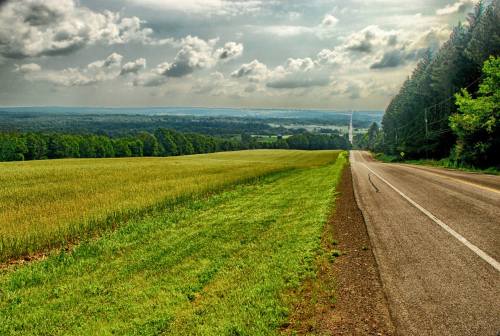 A winding road stretches through lush green fields under a cloudy sky, with distant hills visible in the background.