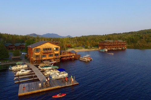 A scenic lakeside view featuring wooden lodges, boats, and a dock surrounded by lush greenery and mountains.