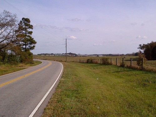 A winding rural road bordered by grass and a fence, with open fields and a cloudy sky in the background.