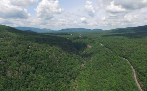 Aerial view of lush green mountains and valleys under a partly cloudy sky. A winding road is visible in the foreground.