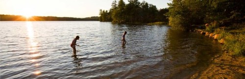 Two children wade in a calm lake at sunset, surrounded by trees and a sandy shore.