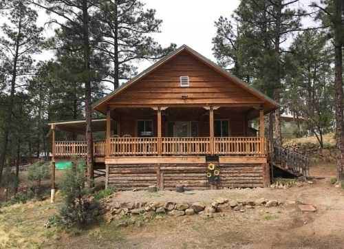 A wooden cabin with a porch, surrounded by tall pine trees and rocky landscaping.