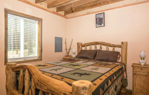 Cozy bedroom with a wooden bed, patterned quilt, and natural light from a window. Rustic decor and warm tones.
