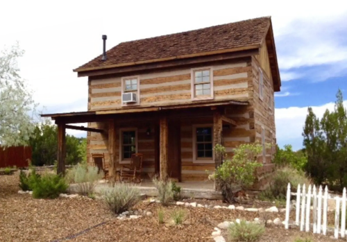 A rustic two-story log cabin with a porch, surrounded by desert landscaping and a white picket fence.
