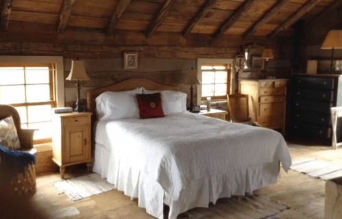 Cozy bedroom with a wooden ceiling, a white bedspread, bedside tables, and rustic decor. Natural light from windows.