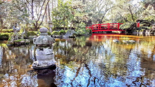 A serene garden scene featuring a stone lantern, a red bridge, and reflections in a tranquil pond surrounded by greenery.