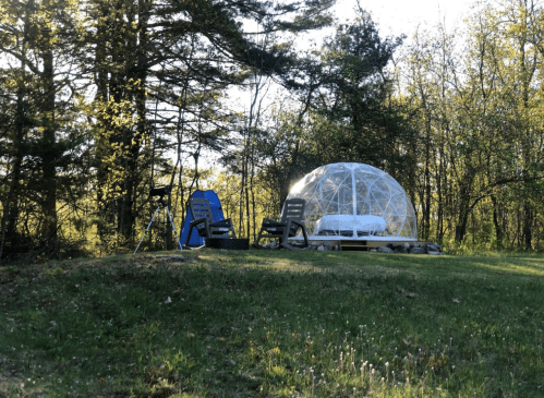 A clear dome structure with a bed, surrounded by trees, and two chairs on a grassy area.