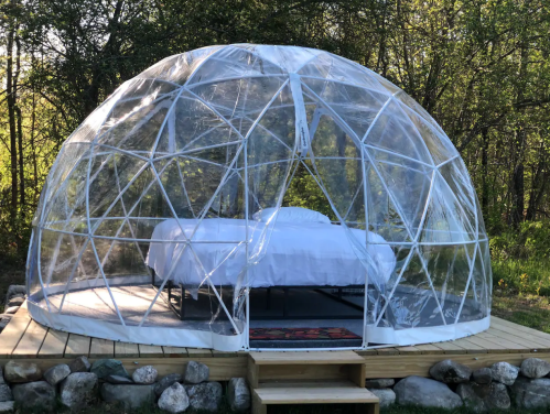 A transparent geodesic dome with a bed inside, set on a wooden platform in a forested area.