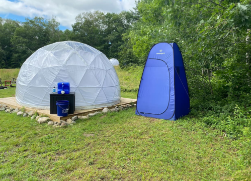 A white geodesic dome and a blue privacy tent on a grassy area surrounded by trees.