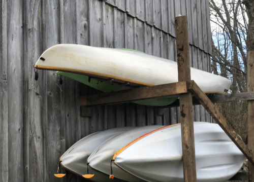Three kayaks stacked on a wooden rack against a weathered gray wall.